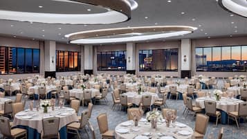 A large banquet hall with round tables set for dining, featuring a cityscape view through large windows.