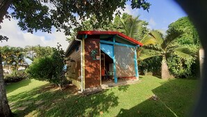 A small, single-story wooden structure with a red roof, surrounded by lush greenery and palm trees. - "LE TI-COLIBRI" CHARMING BUNGALOW at the foot of "La Soufrière" volcano (Saint-Claude)