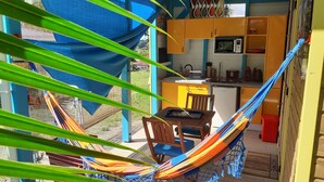 A hammock chair in a sunlit room with a wooden table, a small kitchen area, and a view of the outdoors. - "LE TI-COLIBRI" CHARMING BUNGALOW at the foot of "La Soufrière" volcano (Saint-Claude)