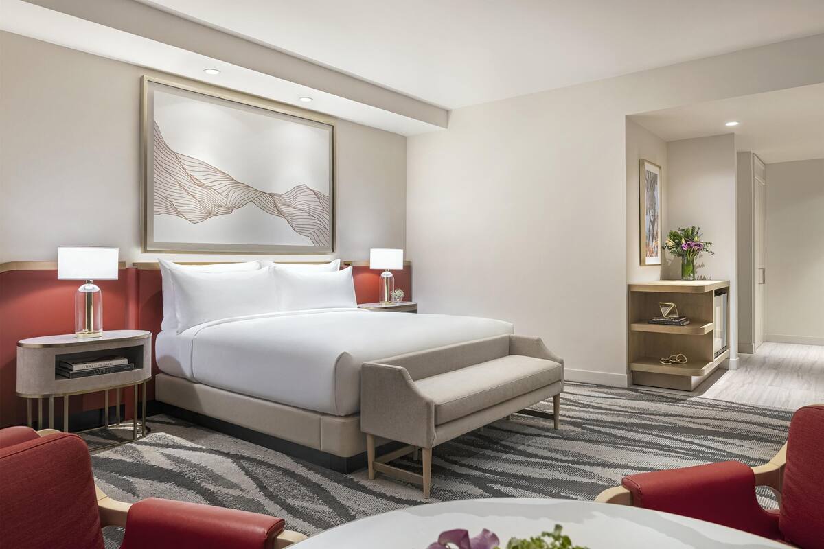 A modern hotel room with a large bed, two bedside tables with lamps, a red armchair, a small table with a vase, and a framed artwork on the wall.
