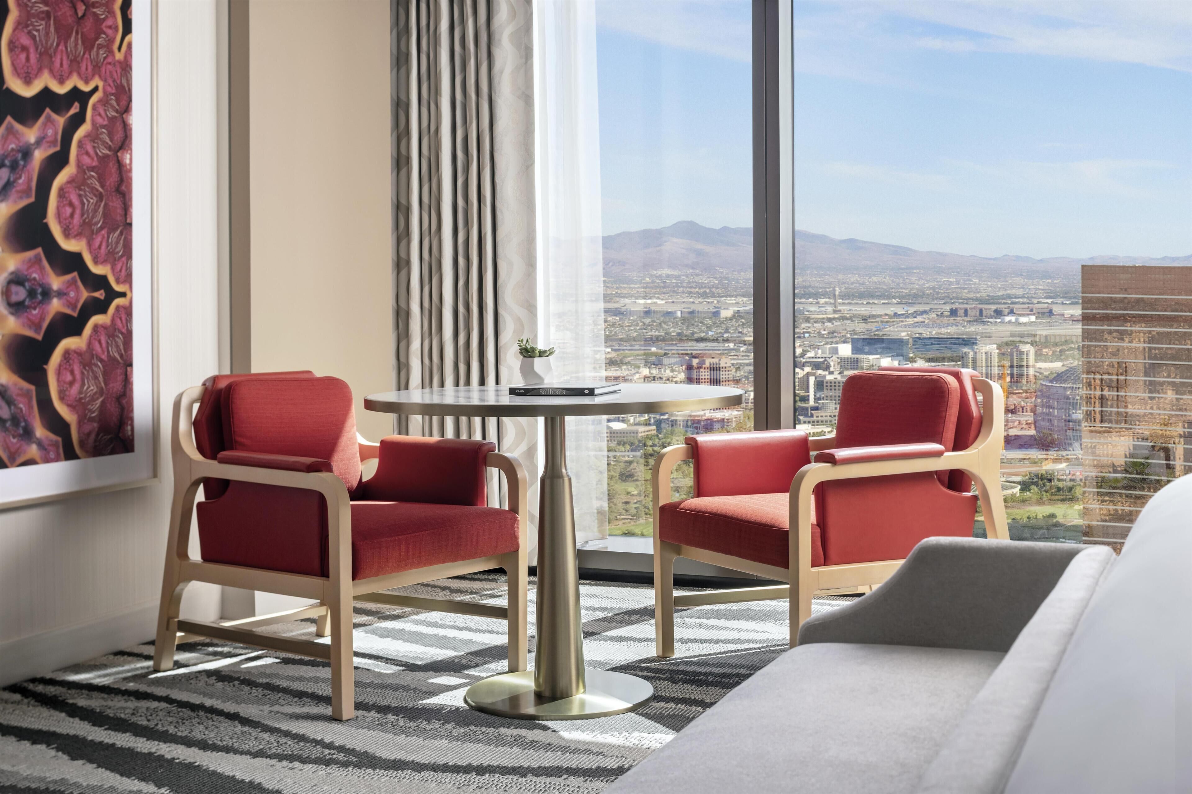 A modern hotel room with a large window offering a city view, two red chairs around a small table, and a grey sofa.