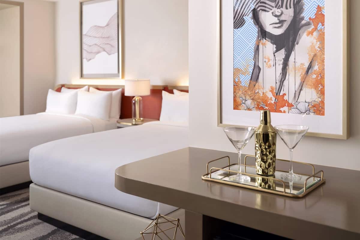 A hotel room with two beds, a bedside table with a bottle and glasses, and a framed artwork on the wall.
