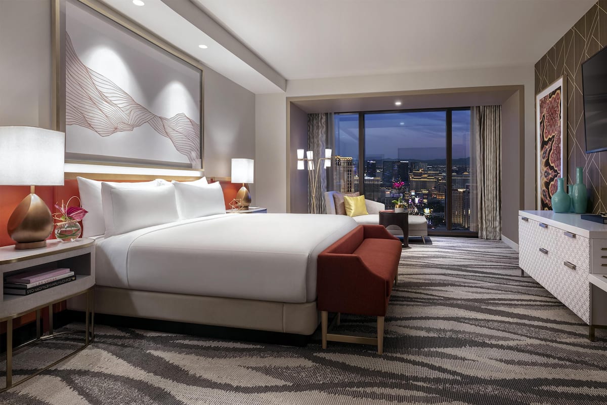 A modern hotel room with a large bed, a seating area, a view of the cityscape, and a large artwork on the wall.