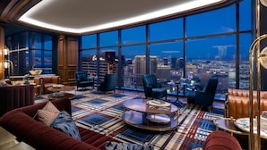 A high-rise hotel room with a large city view, plush seating, and a patterned floor.