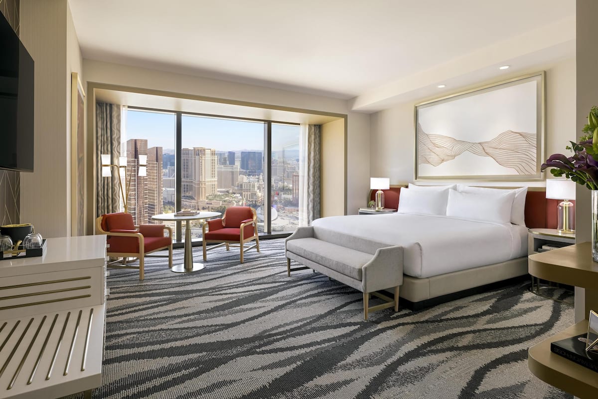 A hotel room with a large bed, a desk, a chair, a small table, and a view of the cityscape through large windows.