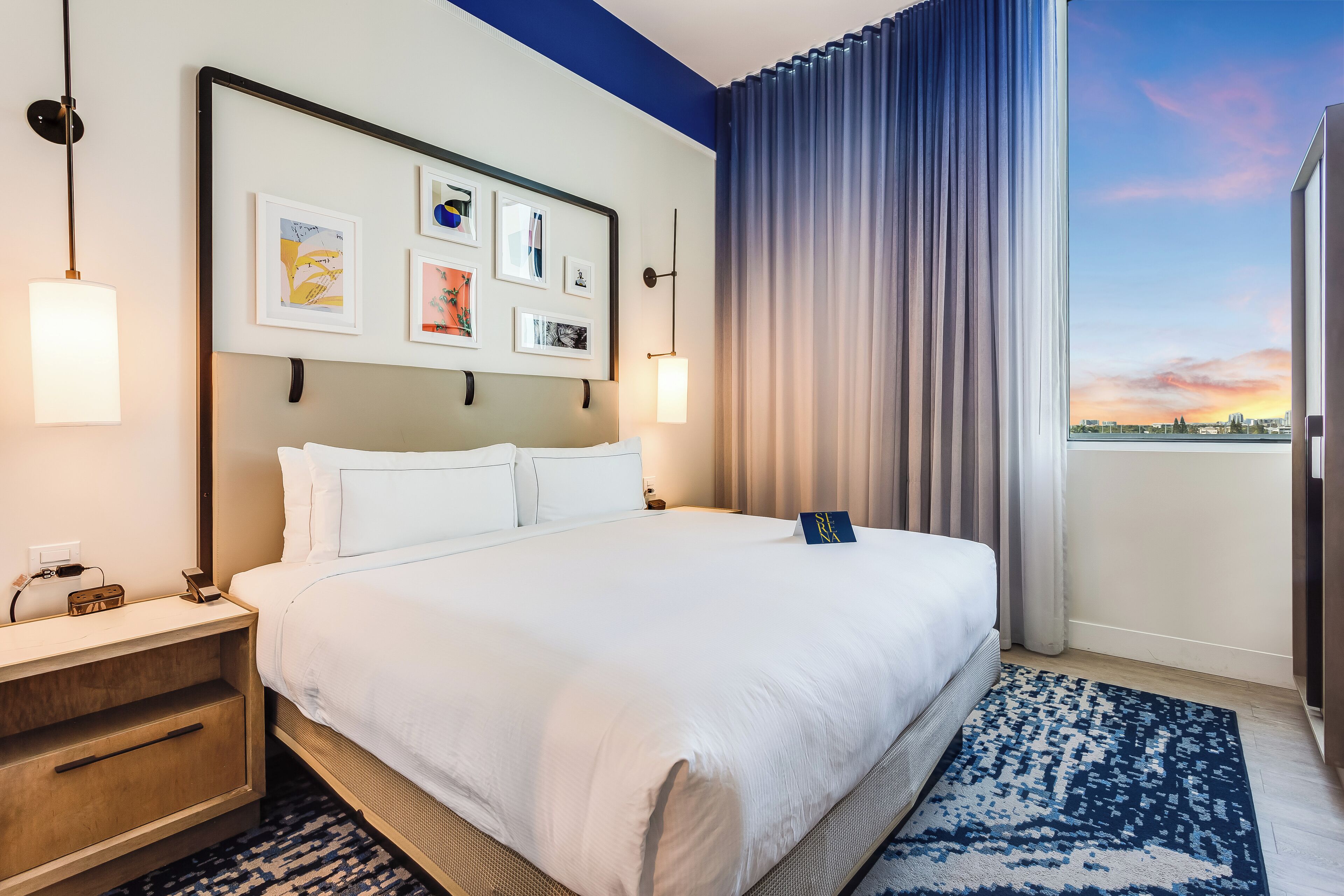 A hotel room with a large bed, a nightstand, a wall-mounted lamp, and a view of the sunset through a window with curtains.