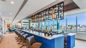 A modern bar area with a long counter, multiple seating options, and a large window offering a city view. - SERENA Hotel Aventura Miami, Tapestry Collection by Hilton (Aventura)