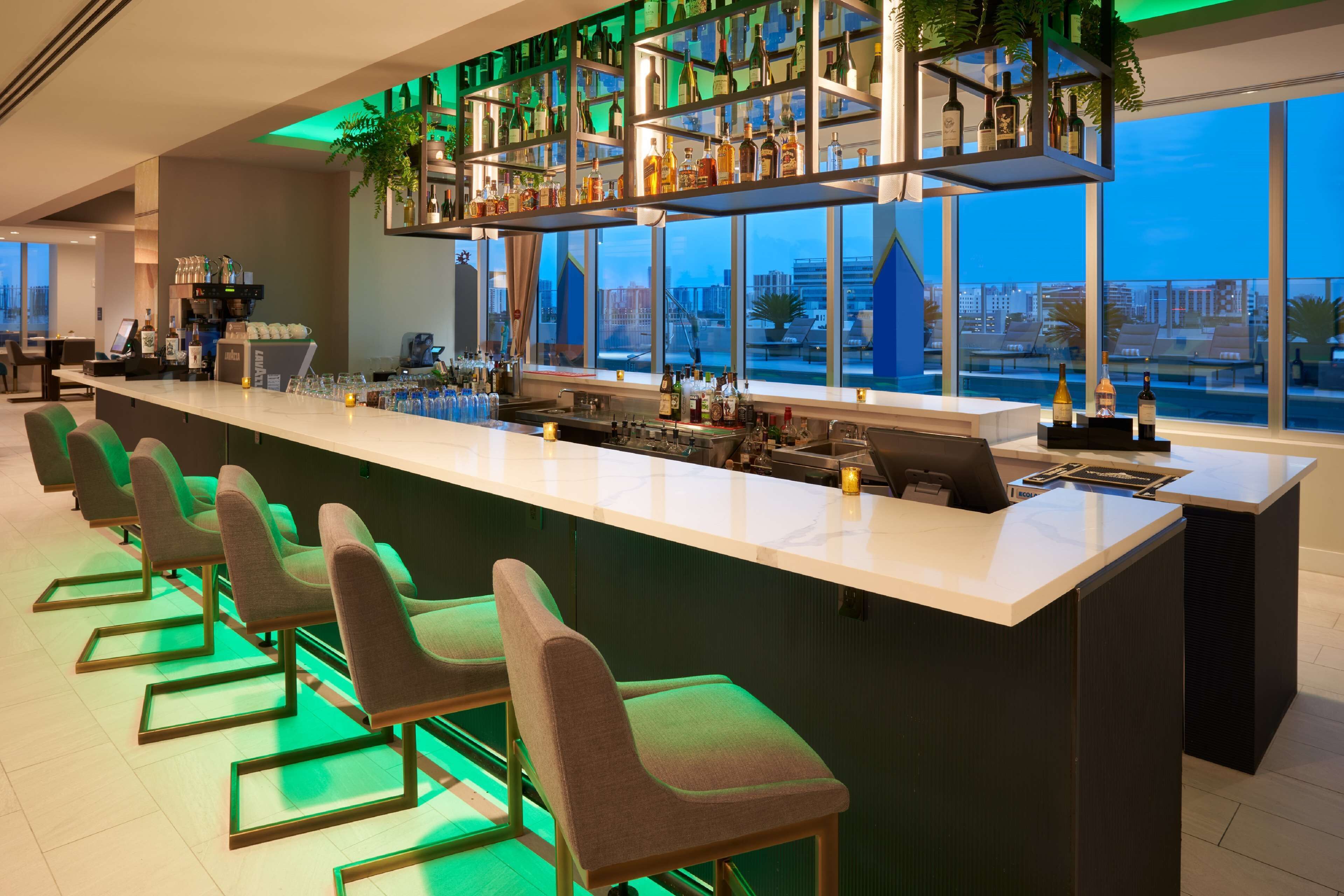 A modern bar area with a long counter, multiple seating options, and a view of the cityscape through large windows.