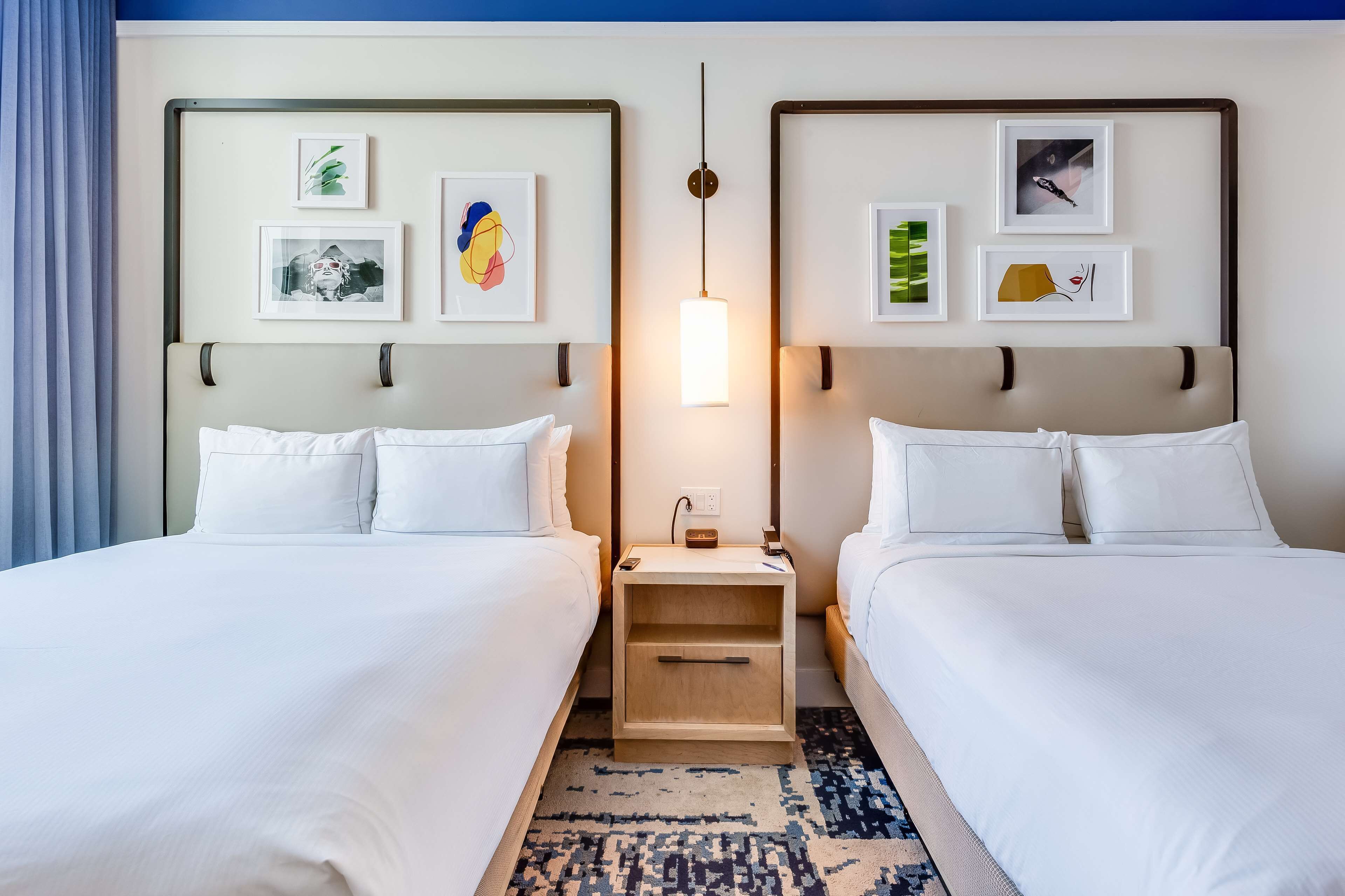 A hotel room with two beds, a nightstand with a lamp, and framed artwork on the walls.