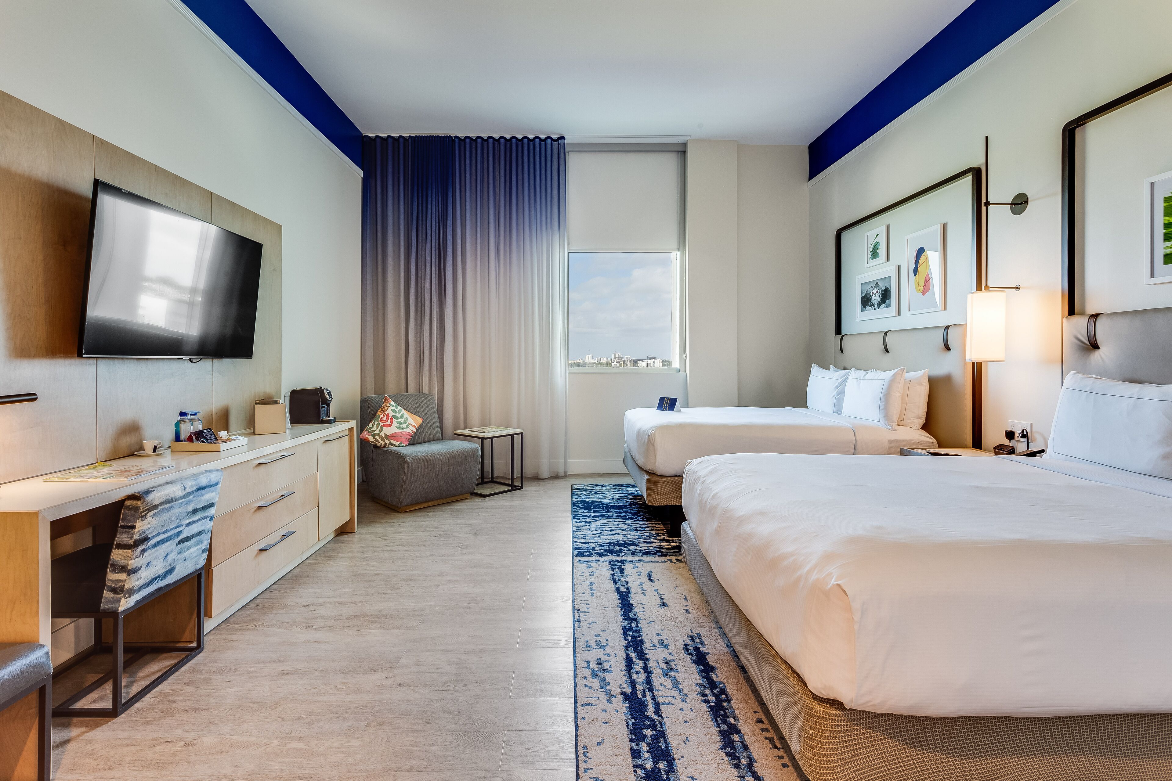 Quarto de hotel com televisão de tela plana, duas camas, uma escrivaninha com cadeira, uma mesa pequena e uma janela com vista.