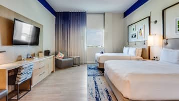 Quarto de hotel com televisão de tela plana, duas camas, uma escrivaninha com cadeira, uma mesa pequena e uma janela com vista.