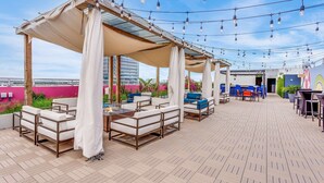 An outdoor patio with white cushioned seating, a wooden pergola, and string lights. - SERENA Hotel Aventura Miami, Tapestry Collection by Hilton (Aventura)