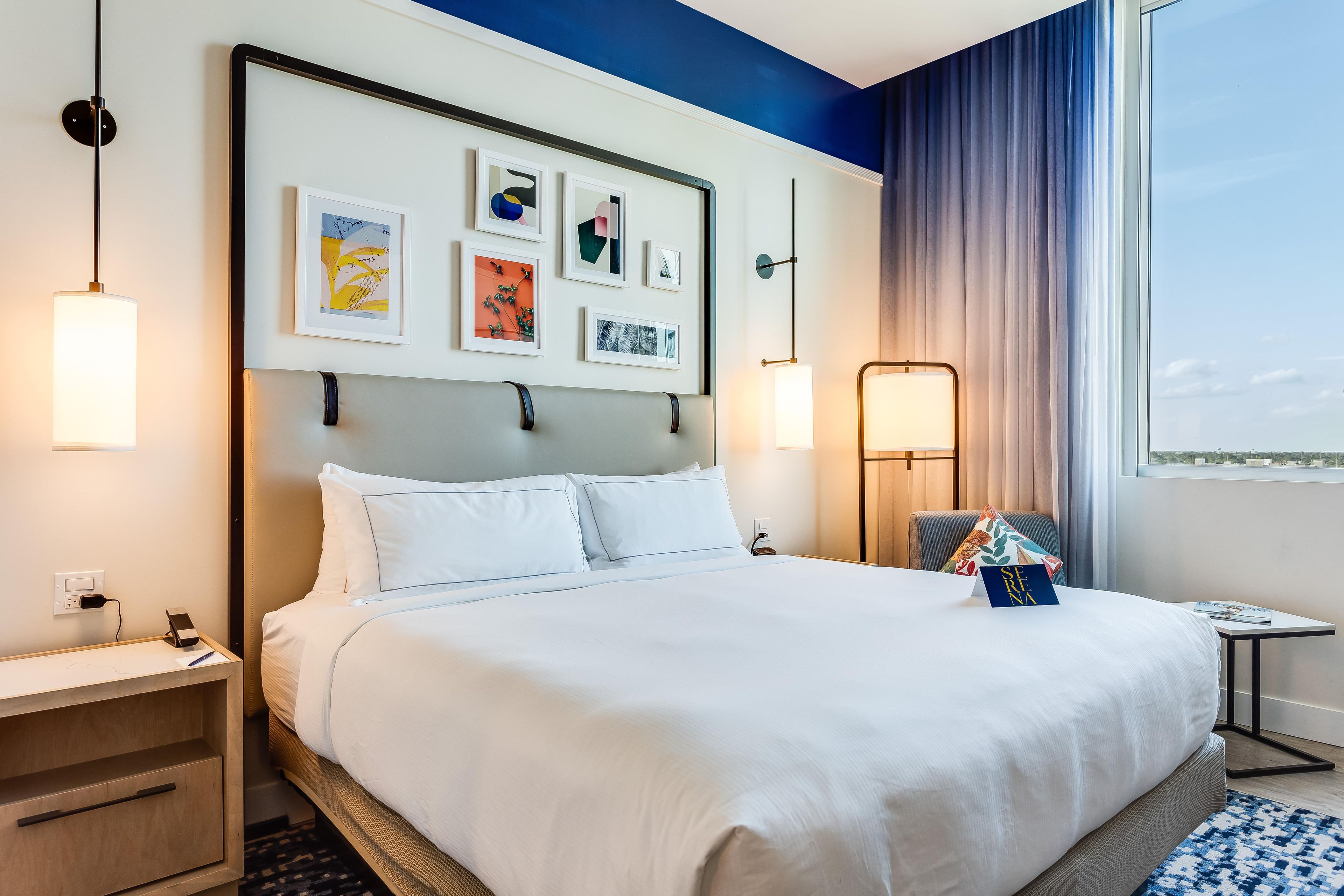 Une chambre d’hôtel avec un grand lit, une table de chevet, une commode, une lampe et une fenêtre avec des rideaux.