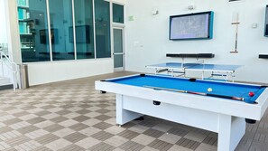 Une table de billard recouverte d’un drap bleu, une table blanche et un queue de billard. Un tableau encadré est accroché au mur et une poubelle se trouve près de la table.