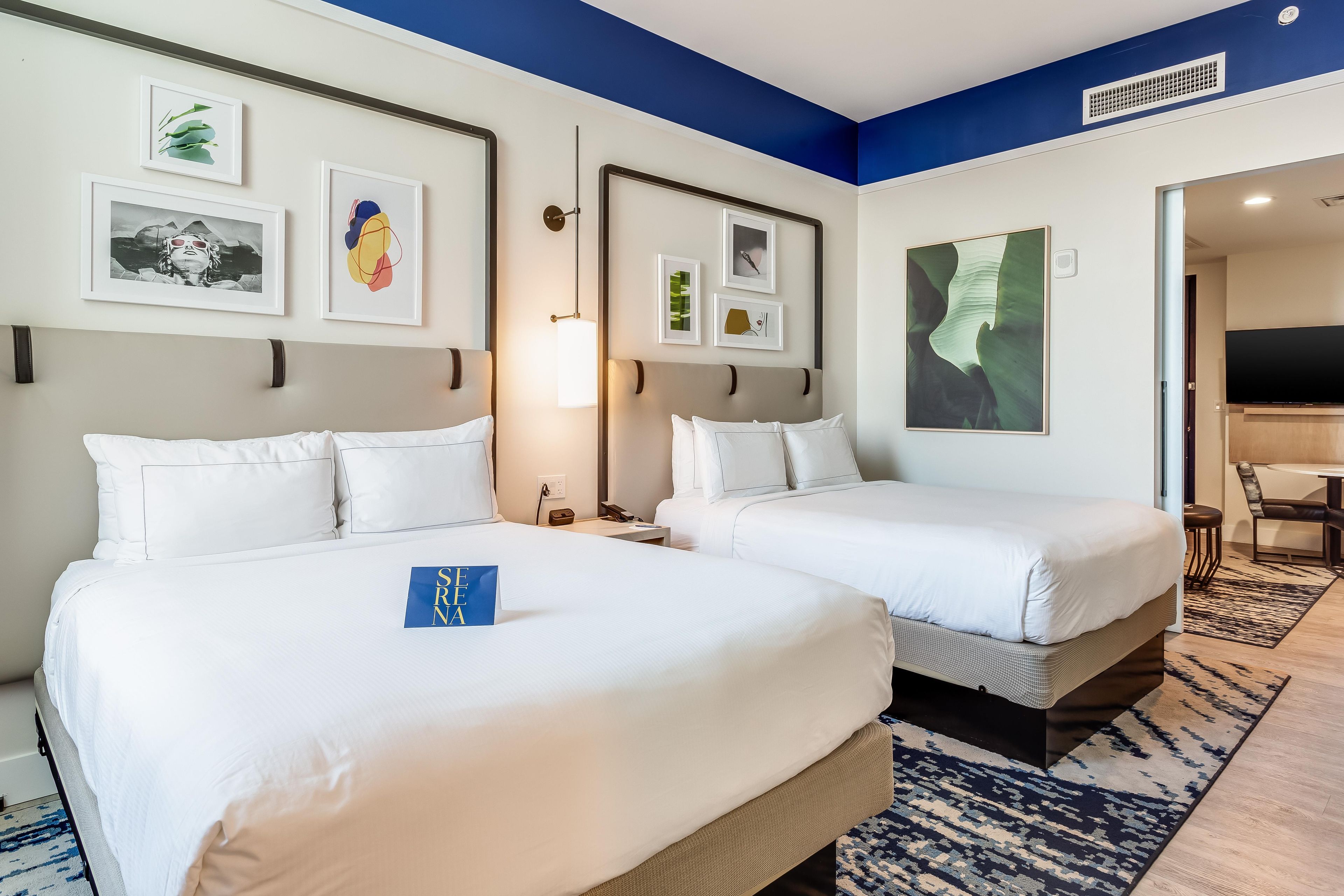A hotel room with two beds, a desk, a chair, and artwork on the walls.