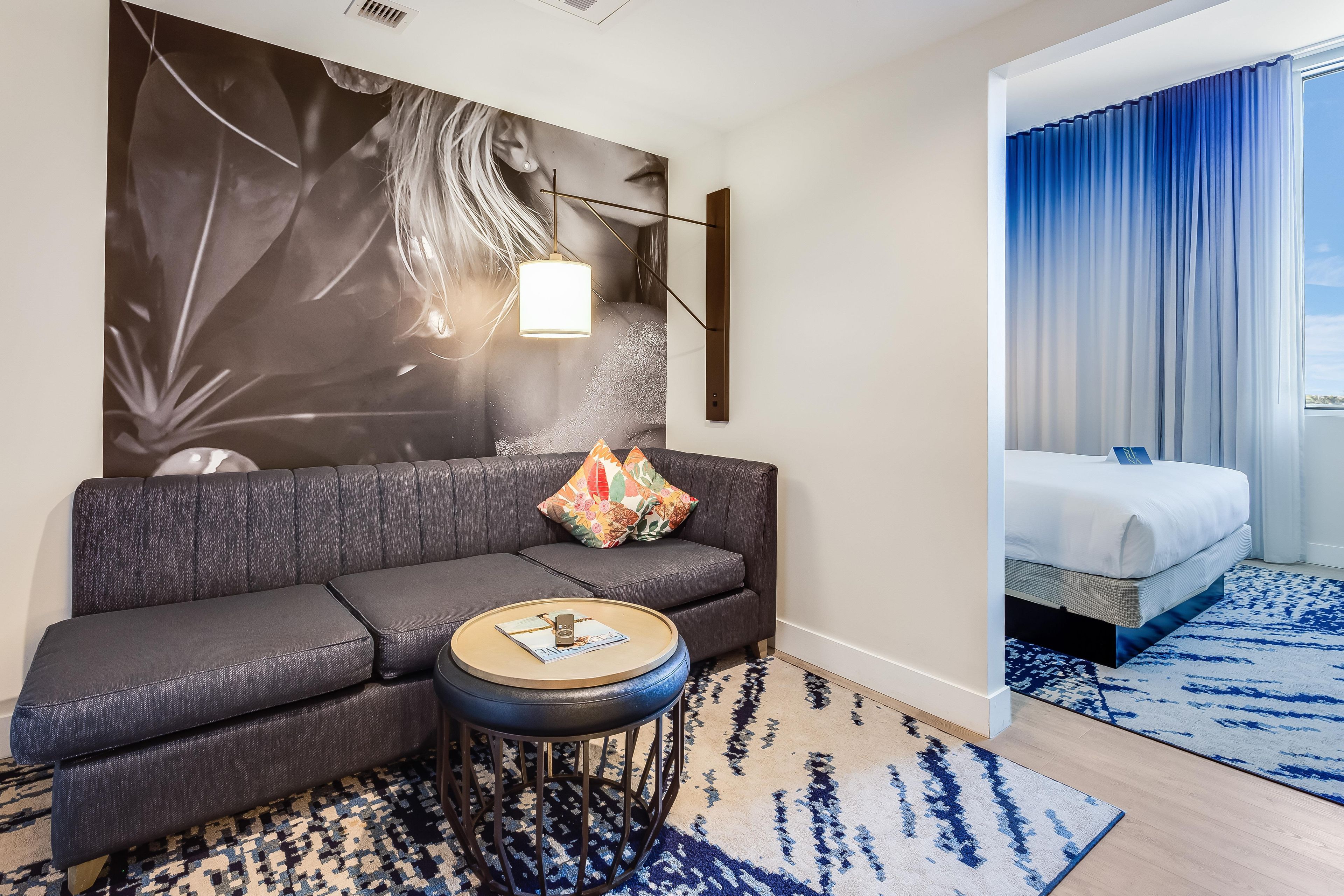 Quarto de hotel com sofá, mesa redonda pequena, parede decorativa, cama com cortina azul e tapete estampado.