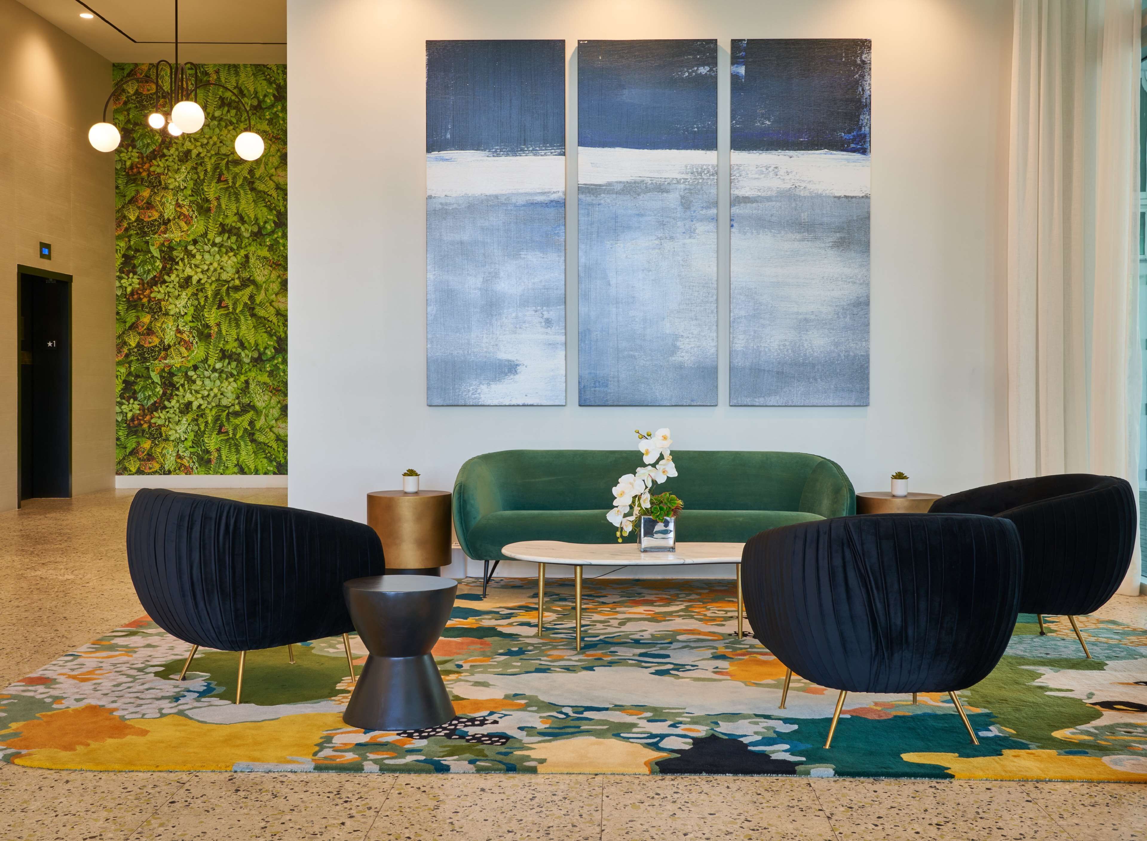 A lobby with a green sofa, two black armchairs, a small table with a vase of flowers, a colorful patterned rug, and a large abstract painting on the wall.