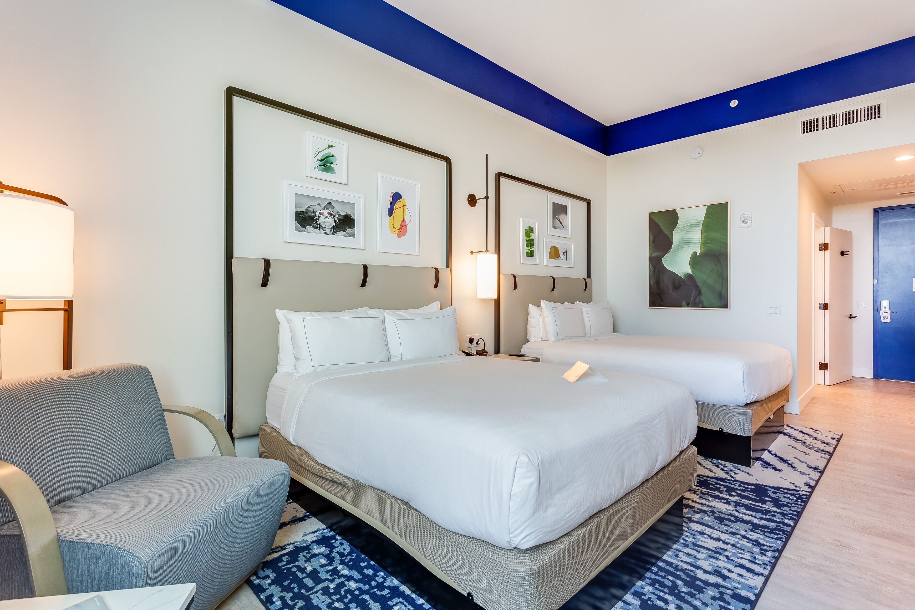 A hotel room with a large bed, a smaller bed, a sofa, and a desk. There are framed pictures on the wall, a lamp, and a door.
