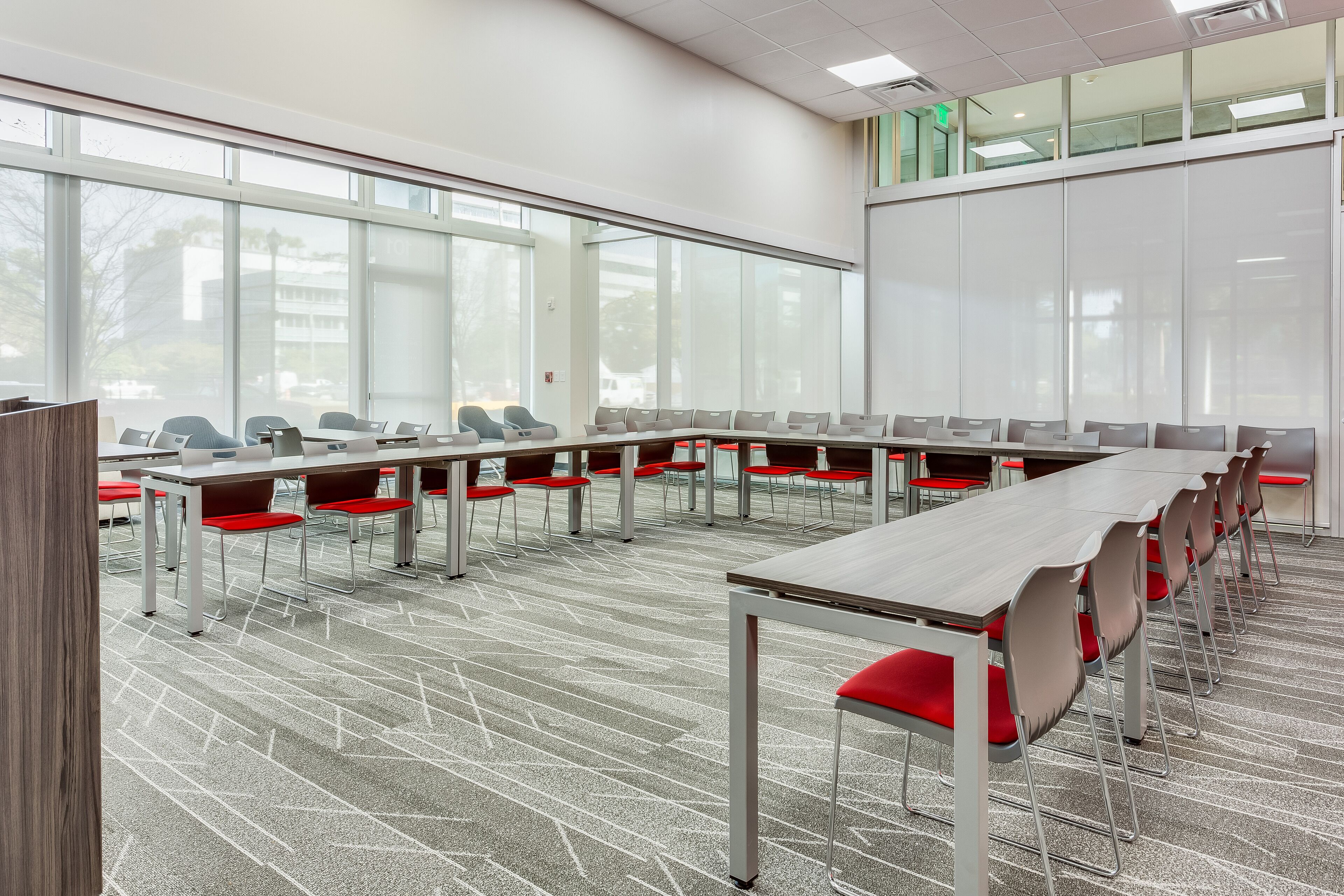 A conference room with long tables and red chairs, large windows, and a carpeted floor.
