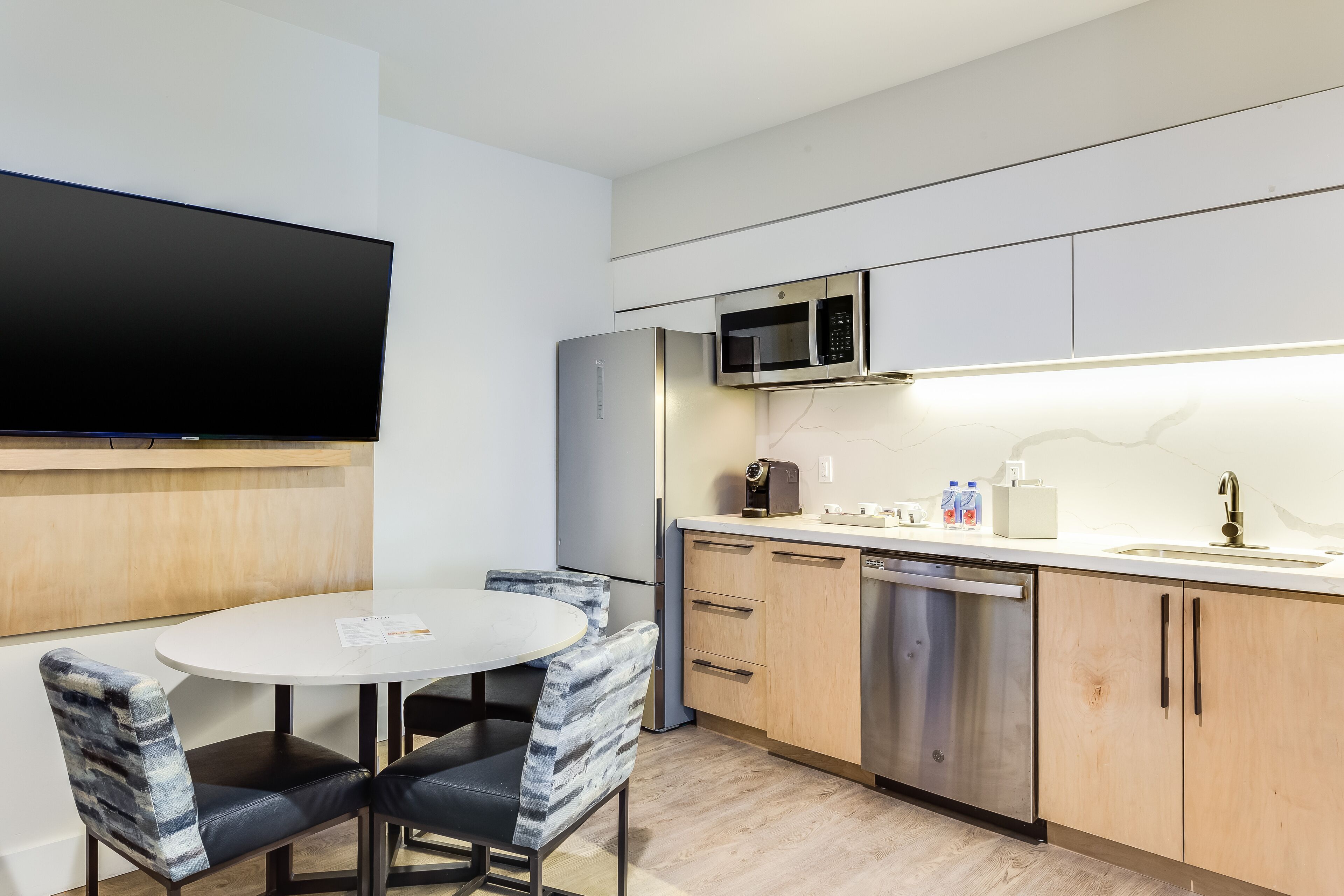 Cocina compacta con microondas, refrigerador y lavavajillas. Hay una mesa redonda con dos sillas y un televisor de pantalla plana montado en la pared.