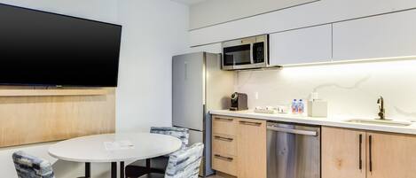 Cocina compacta con microondas, refrigerador y lavavajillas. Hay una mesa redonda con dos sillas y un televisor de pantalla plana montado en la pared.