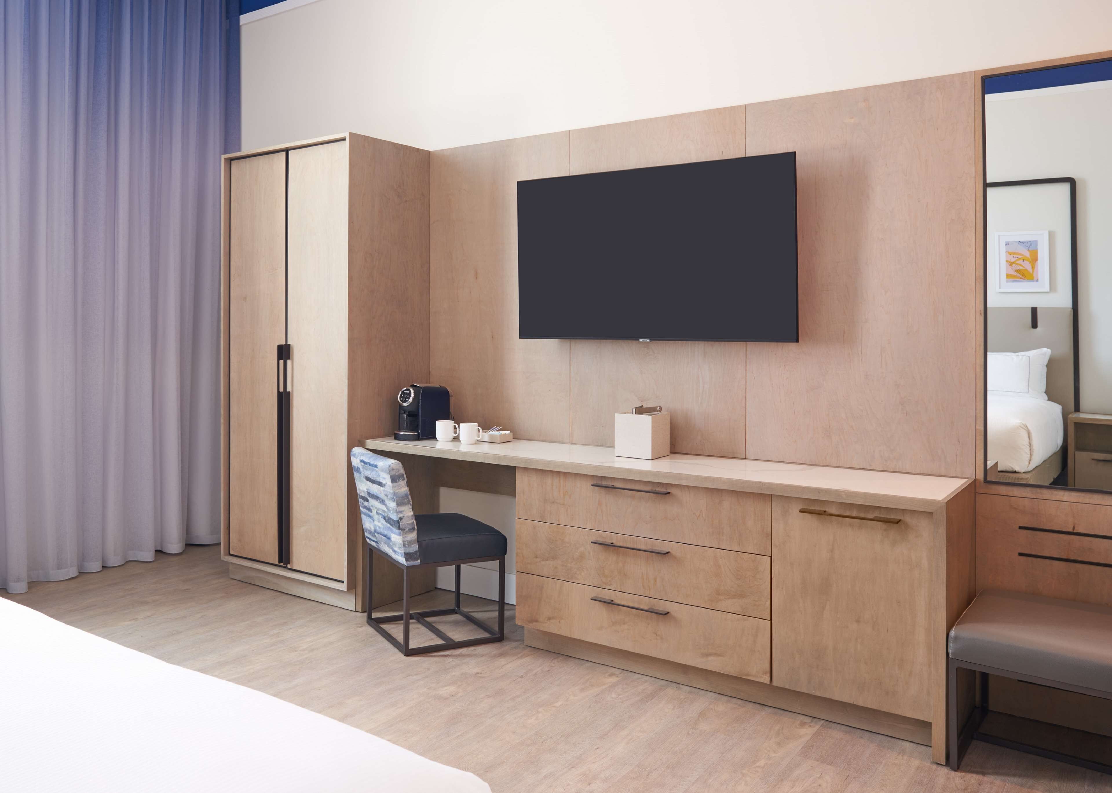 Quarto de hotel moderno com escrivaninha de madeira, uma cadeira, TV de tela plana na parede e uma cama com cabeceira.