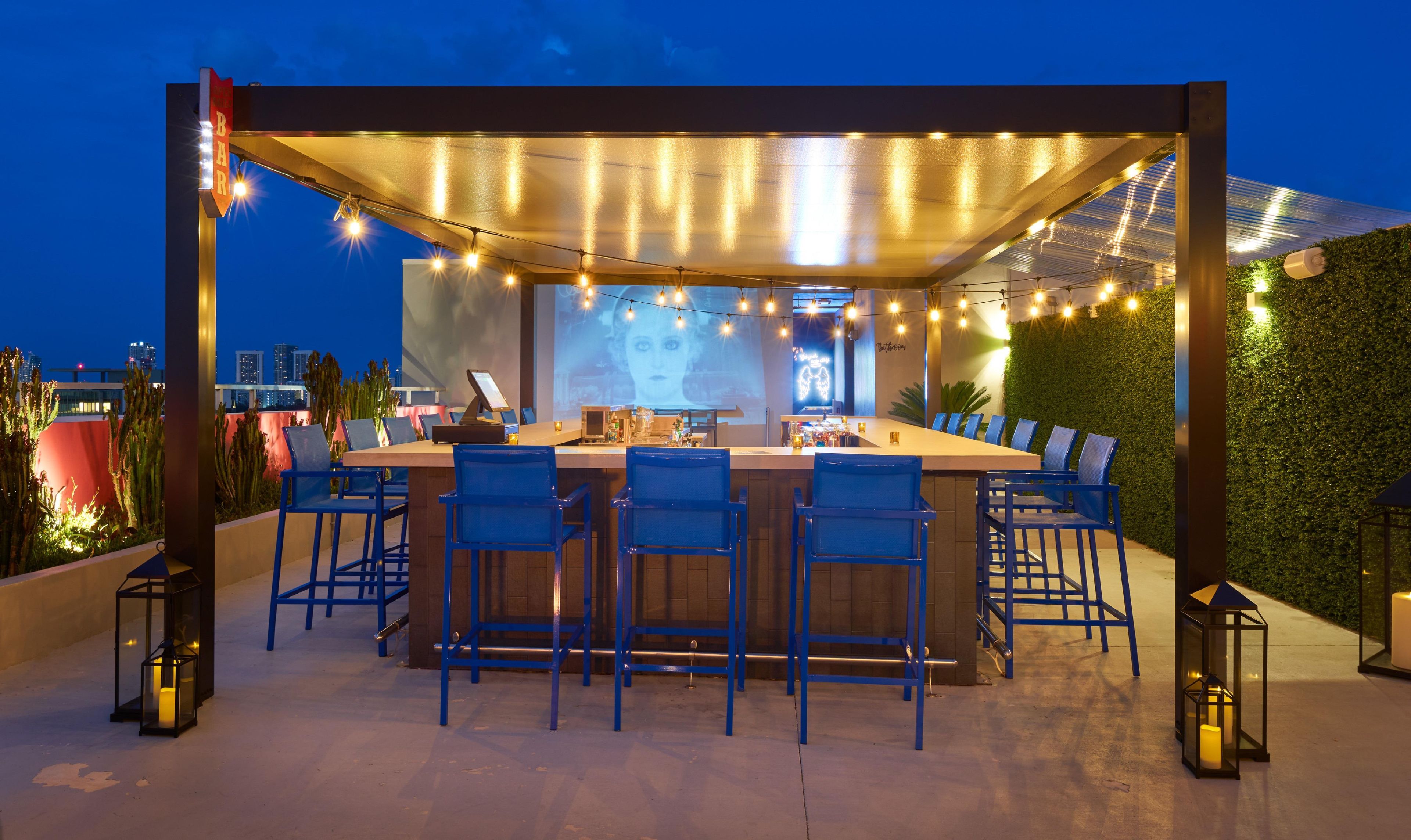 Zona de bar al aire libre con una mesa larga y sillas azules, rodeada de luces colgantes y una pantalla de proyección.