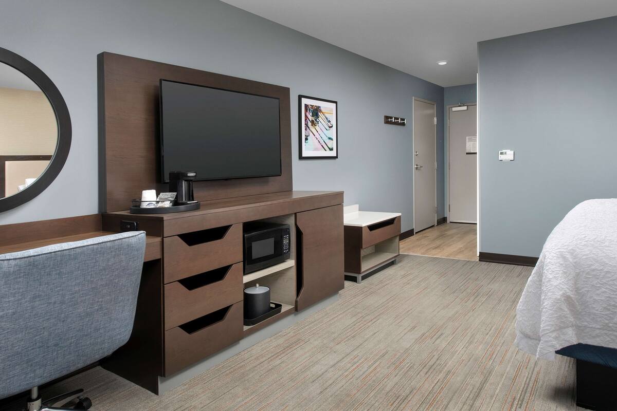 Ein Hotelzimmer mit Flachbildfernseher, Kaffeemaschine, Mikrowelle, Schreibtisch mit Schubladen, Stuhl, Bett und einem gerahmten Bild an der Wand.