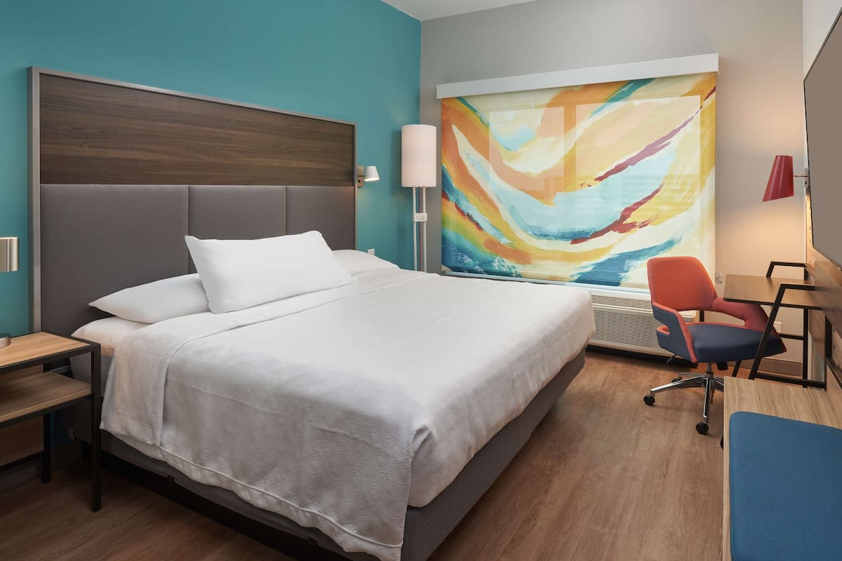 A modern hotel room with a large bed, a desk, and a colorful abstract painting on the wall.