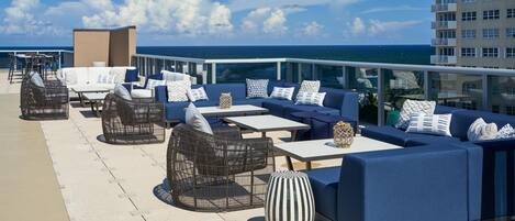 A rooftop bar with outdoor seating, including sofas and chairs, overlooking the ocean.