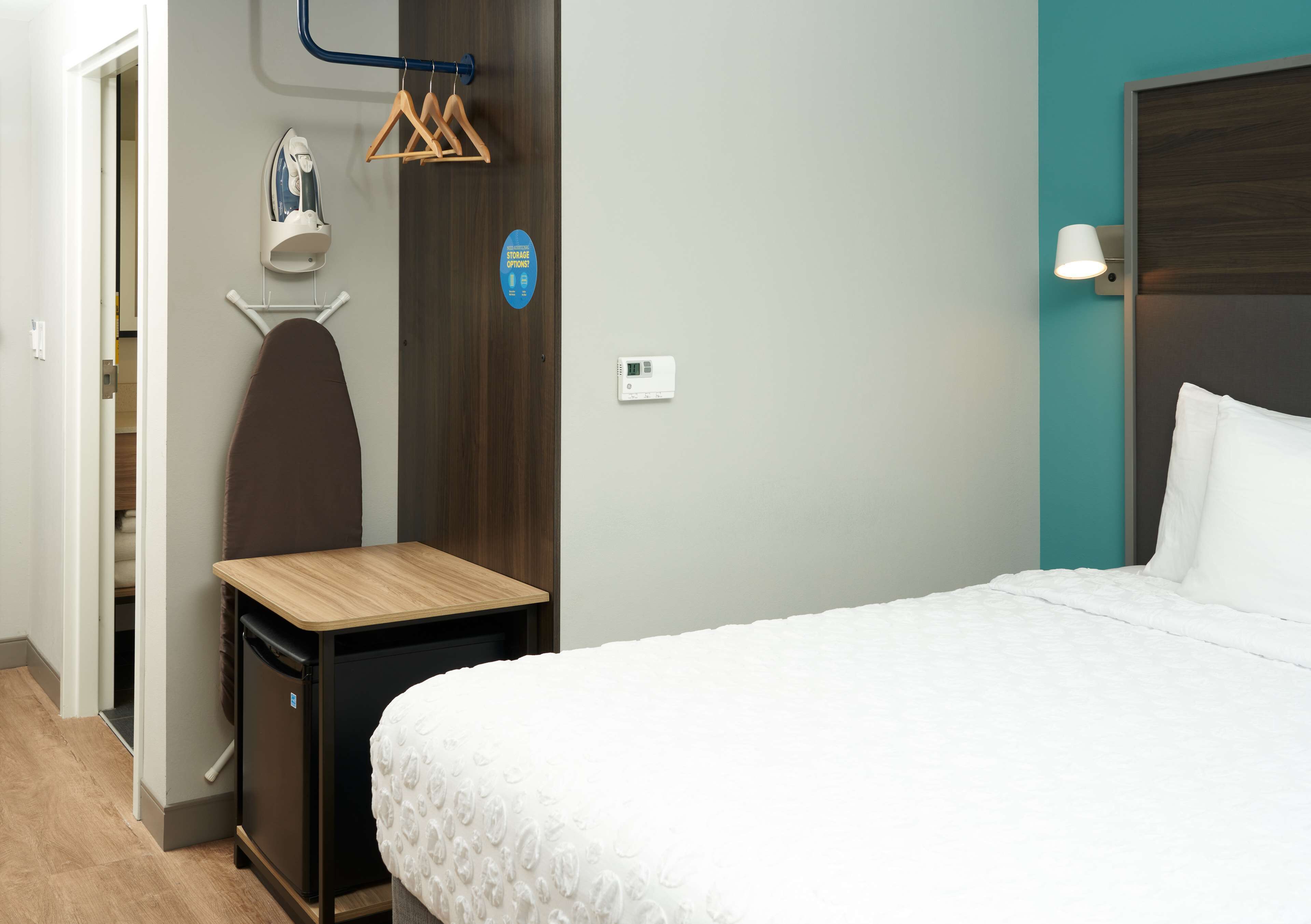 Habitación de hotel con una cama, un escritorio pequeño con silla, una tabla de planchar en la pared, un perchero y un termostato.