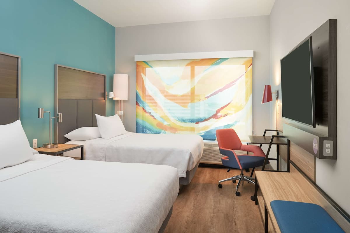 A hotel room with two beds, a desk, a chair, a TV, and a colorful abstract painting on the wall.