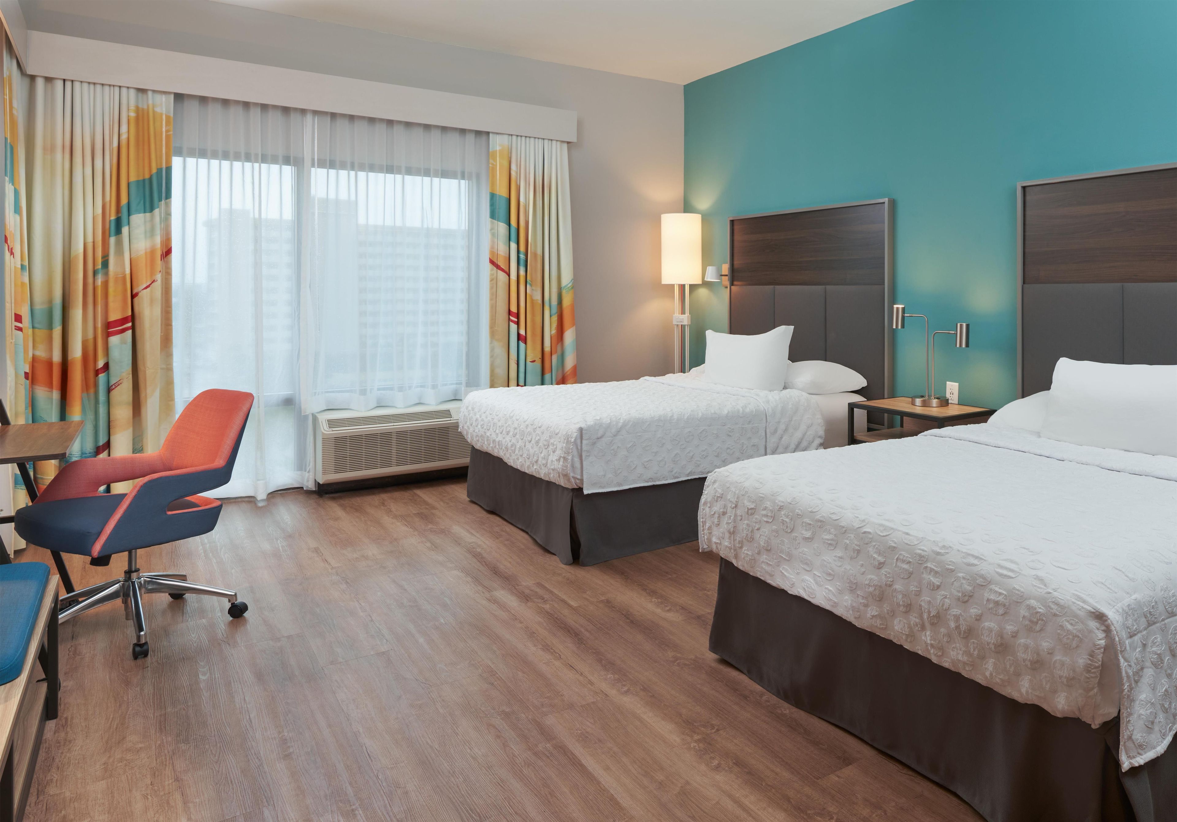A hotel room with two beds, a desk, a chair, a lamp, and a window with curtains.