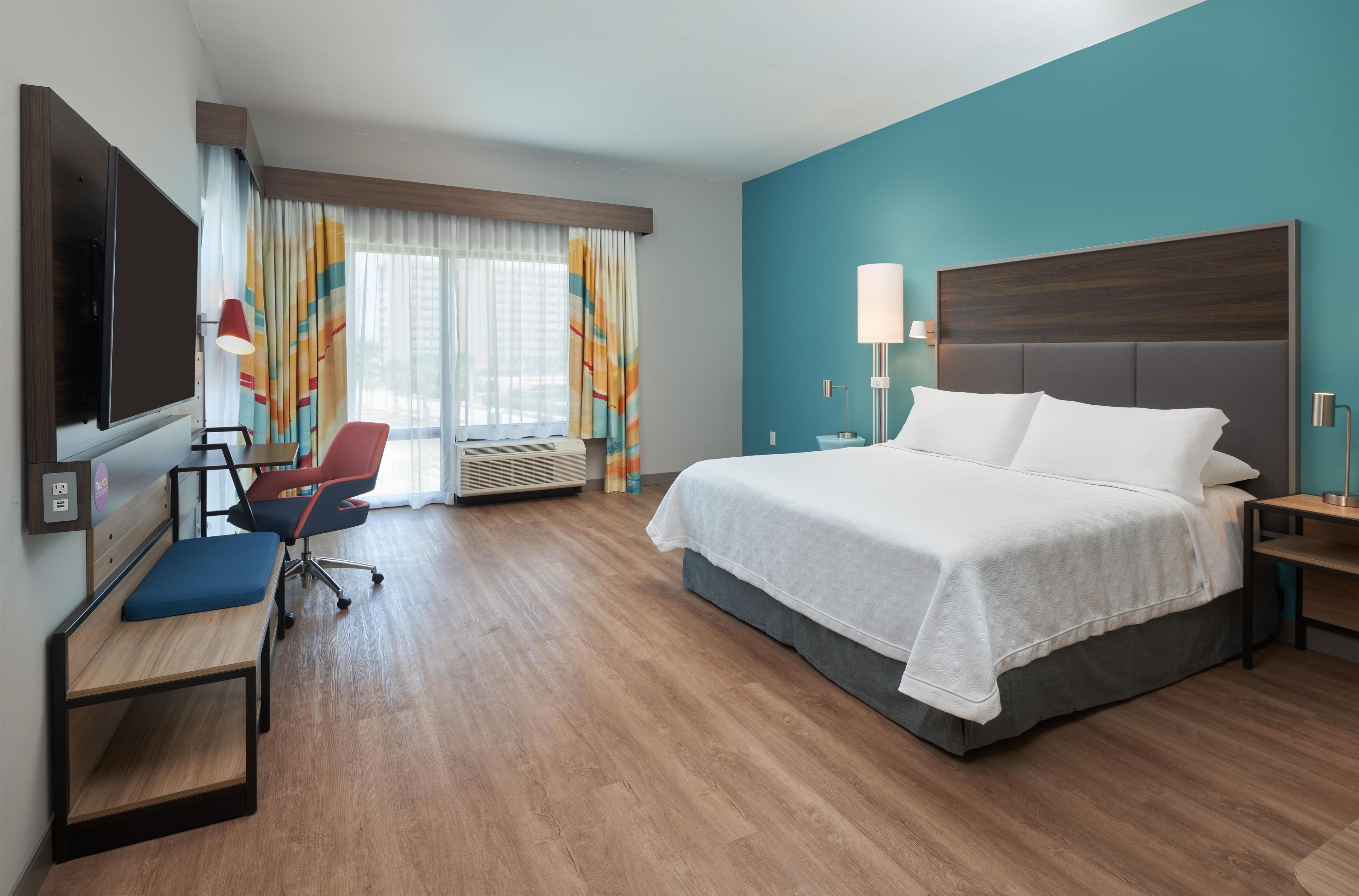 A hotel room with a bed, desk, chair, TV, and a window with curtains.