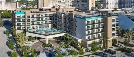 Un hotel de varios pisos con piscina en la azotea, rodeado de palmeras y un estacionamiento.