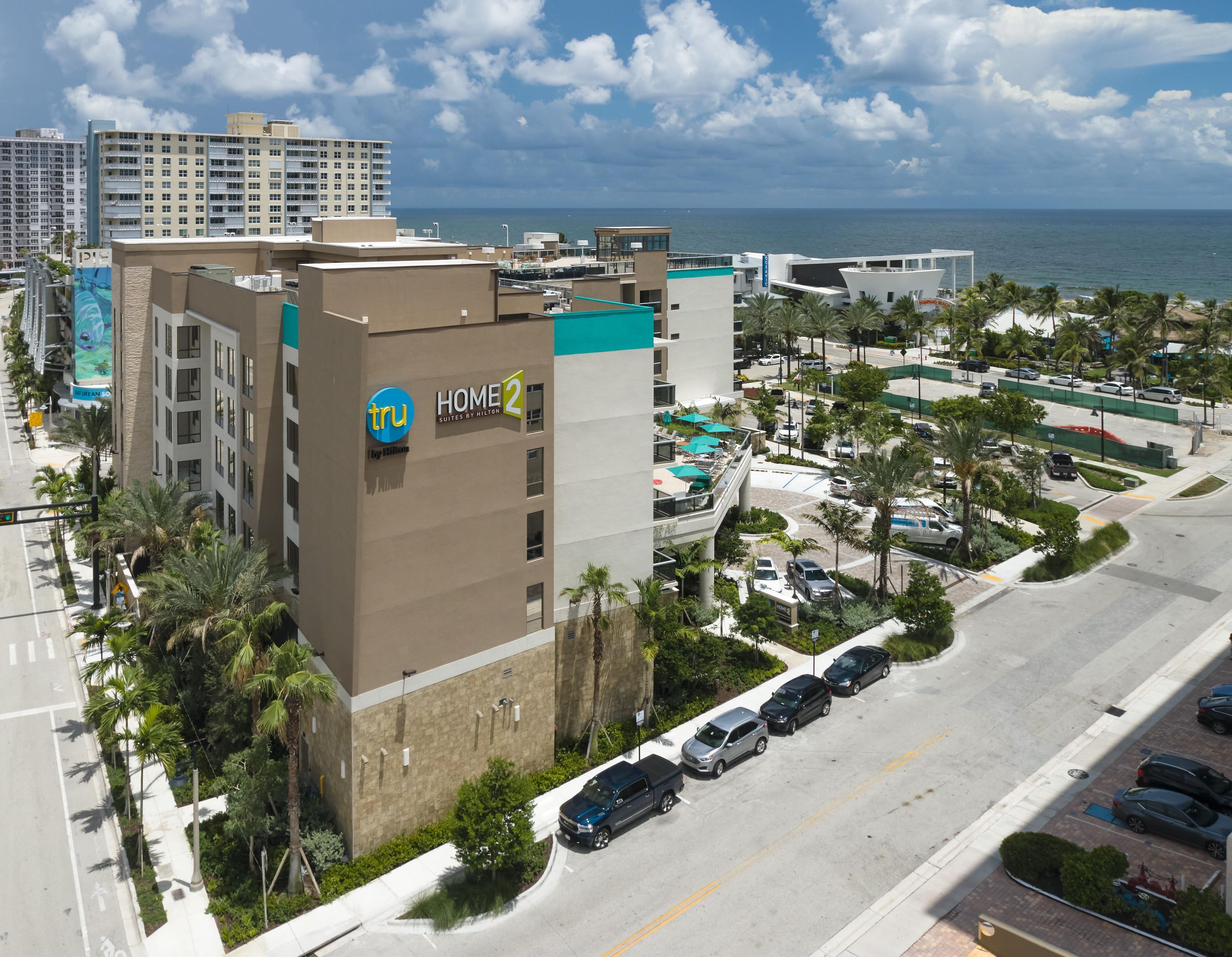 Il s'agit d'un hôtel dont l'enseigne indique « HOME 2 » et « TRU by Hilton », ce qui signifie qu'il fait partie du groupe Hilton. On peut voir plusieurs étages et l'hôtel est entouré d'un parking où sont garées plusieurs voitures.