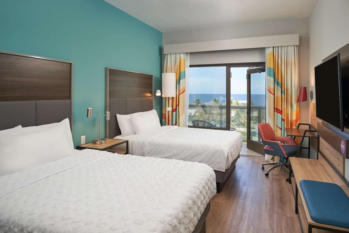 A hotel room with two beds, a desk, a chair, a TV, and a balcony with a view of the ocean.