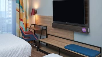 Una habitación de hotel con una cama, un escritorio, una silla, un banco y un televisor instalado en la pared.