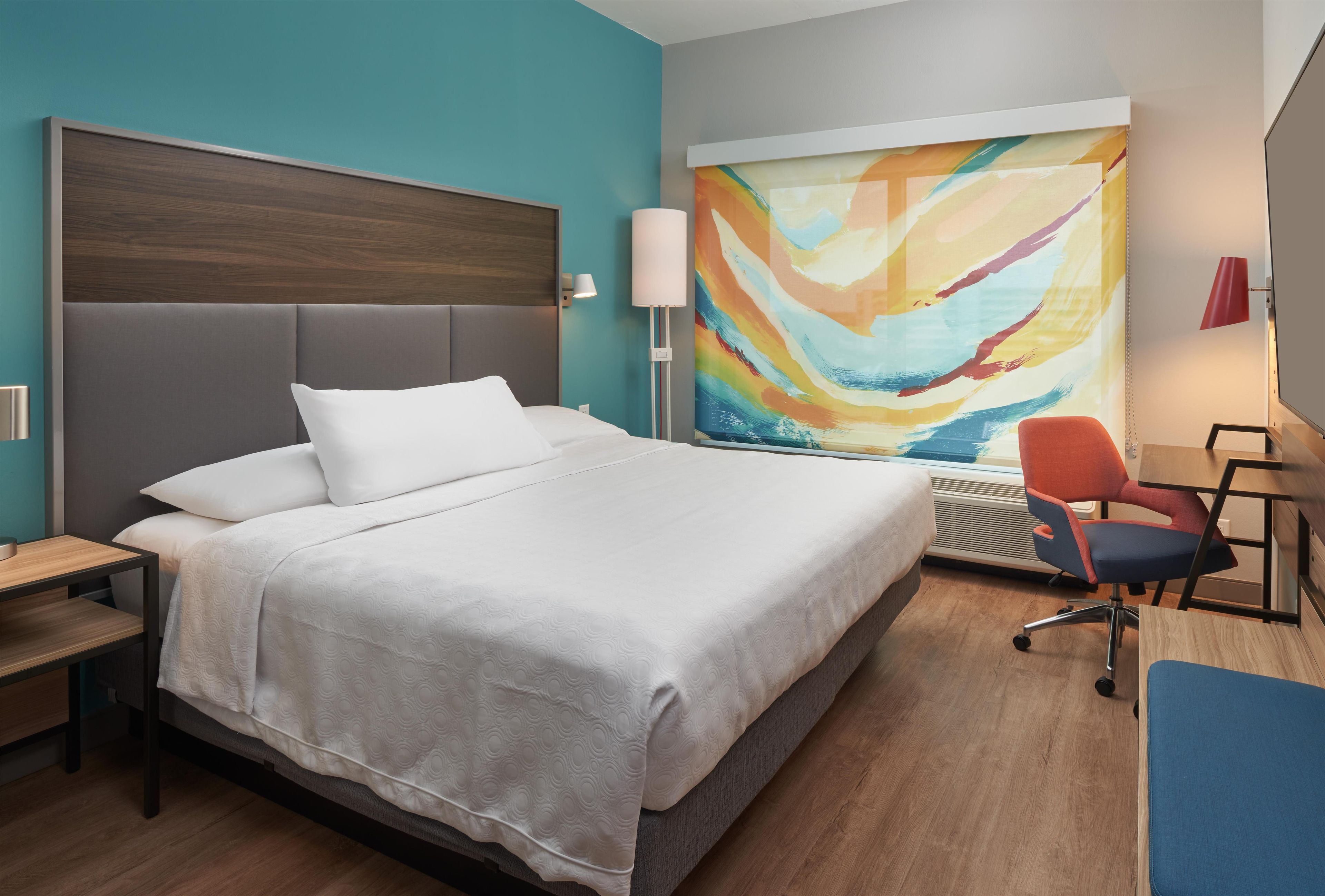 Een hotelkamer met een groot bed, een bureau met stoel, een wandlamp en een kleurrijk abstract schilderij.