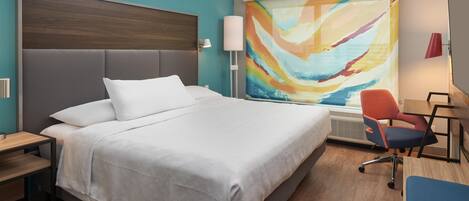 Een hotelkamer met een groot bed, een bureau met stoel, een wandlamp en een kleurrijk abstract schilderij.