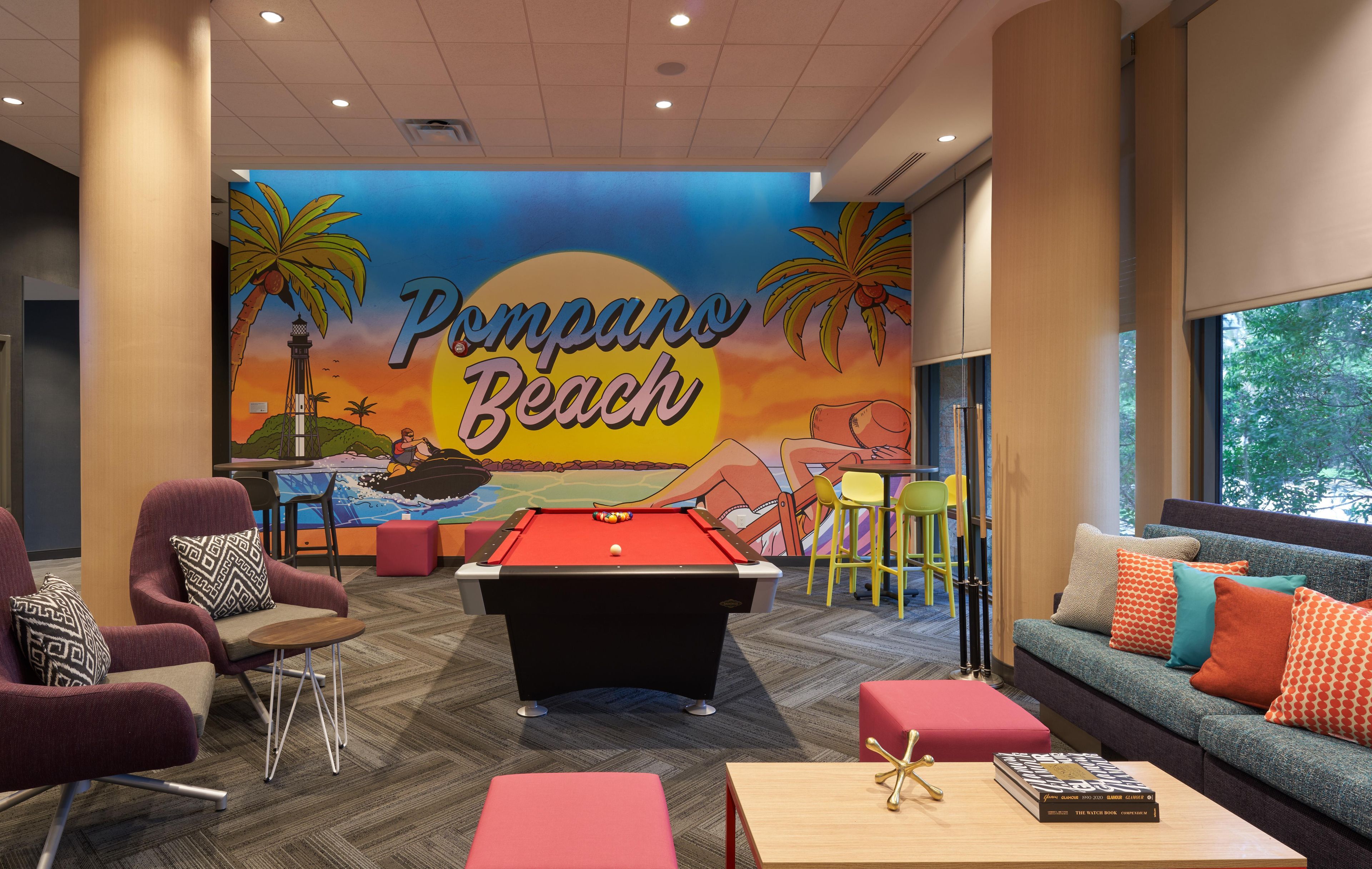 A pool table in a room with a mural of a beach scene, a sofa with colorful cushions, and a book on a table.