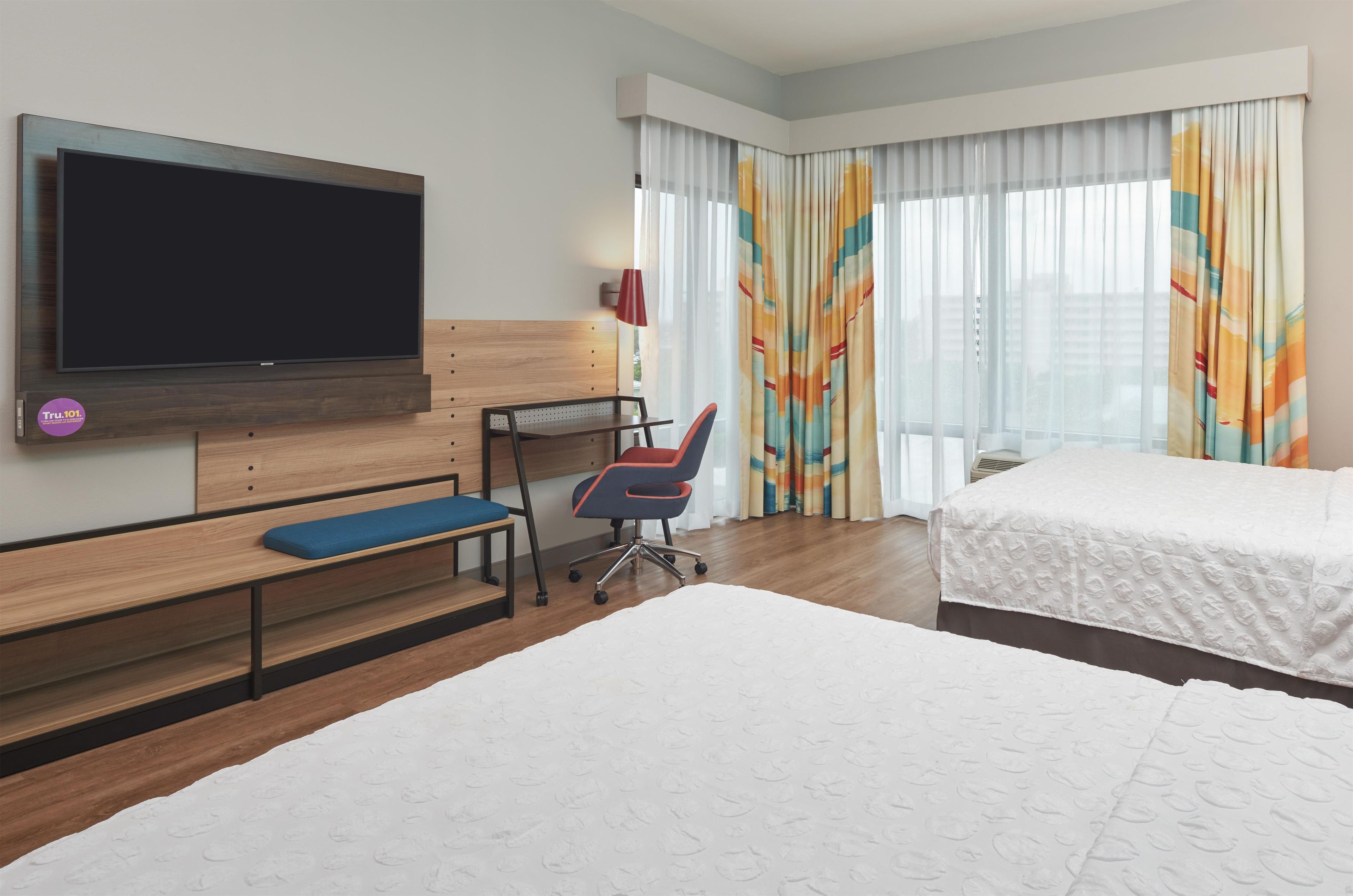 Una habitación de hotel con televisión, escritorio, silla, dos camas y cortinas.