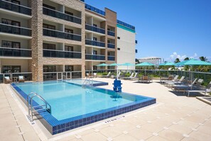 A swimming pool with a blue tiled edge, surrounded by lounge chairs and umbrellas, with a multi-story building in the background. - Home2 Suites by Hilton Pompano Beach Pier (Pompano Beach)
