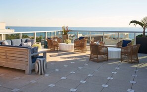 A rooftop terrace with wicker furniture, a round table, and a view of the ocean. - Home2 Suites by Hilton Pompano Beach Pier (Pompano Beach)