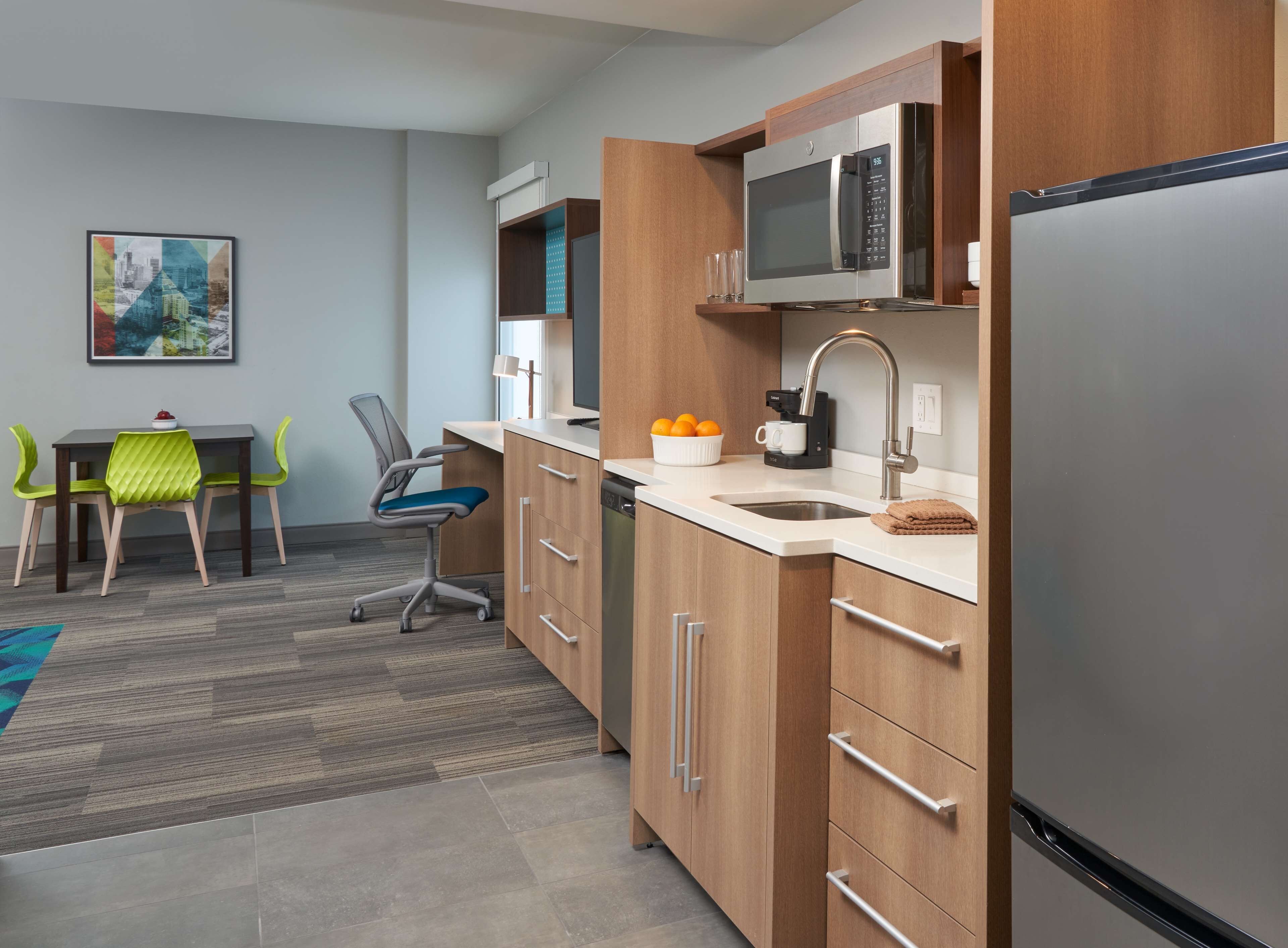 A modern kitchen with wooden cabinets, stainless steel appliances, a dining area with a table and chairs, and a cityscape painting on the wall.