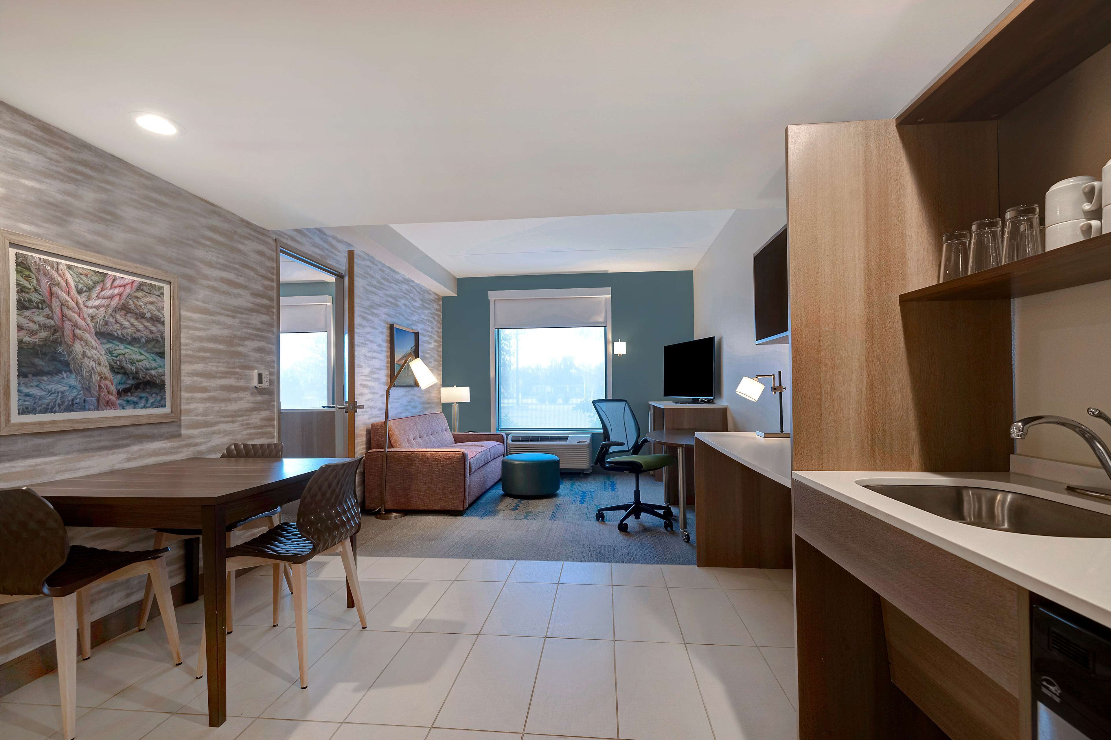 A modern hotel room with a dining area, kitchenette, and a large window providing a view.