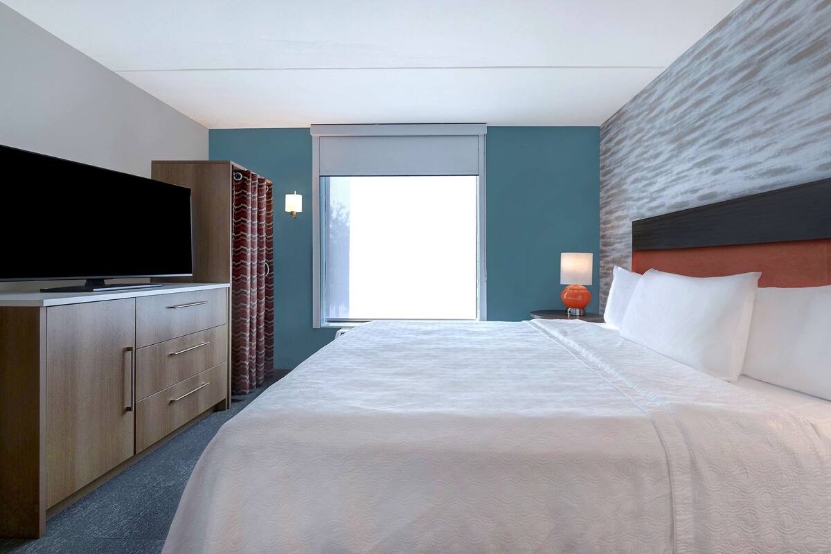 Ett hotellrum med en stor säng, en platt-TV, en byrå i trä och ett fönster med gardiner.