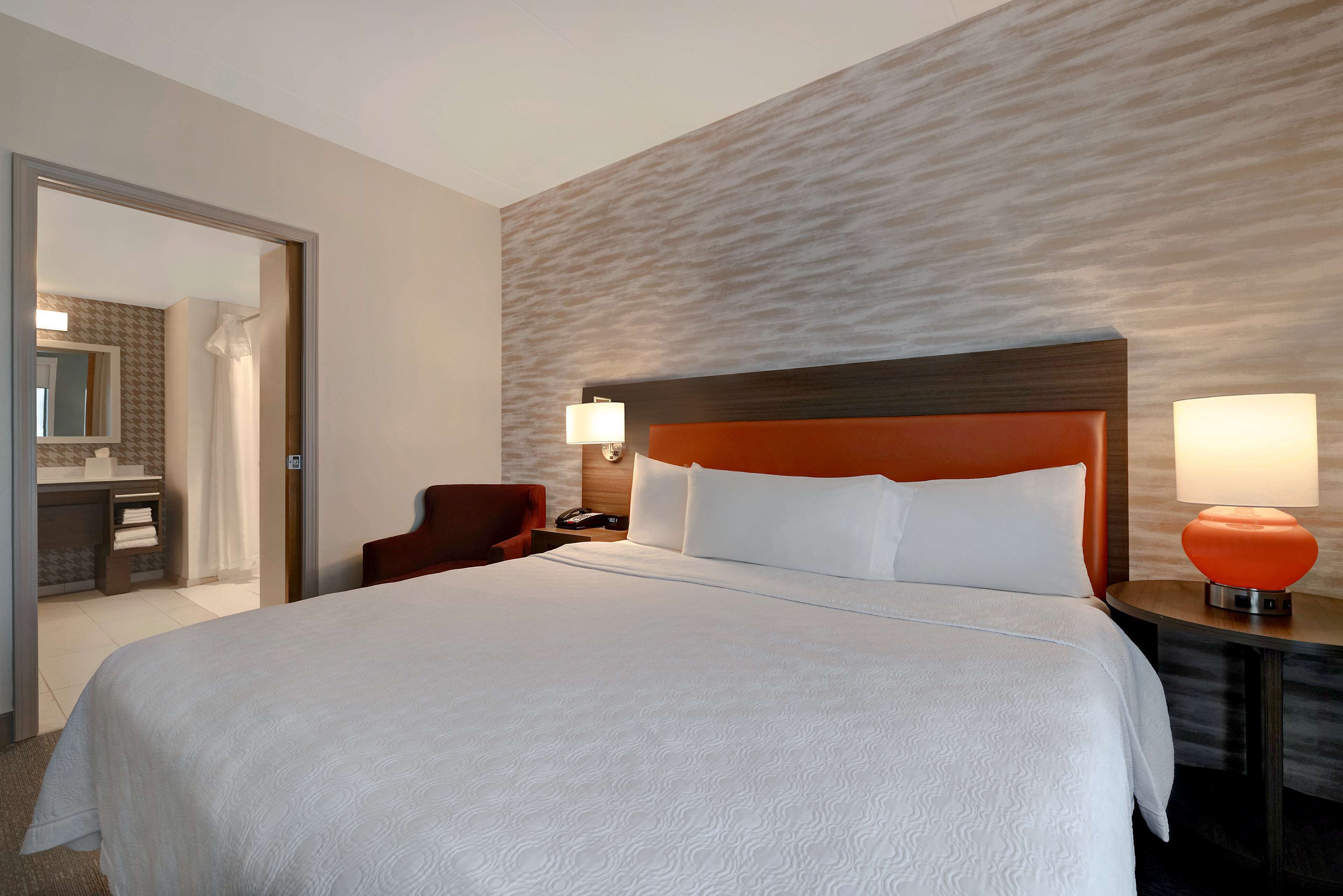 A hotel room with a large bed, a wooden headboard, two bedside lamps, a chair, and a bathroom visible through an open door.