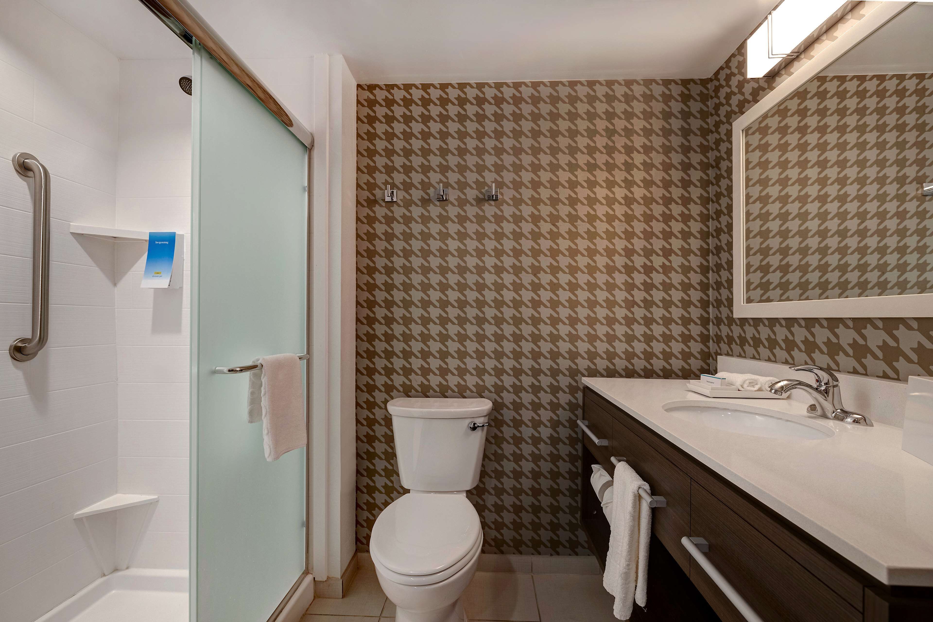 Une salle de bain avec des toilettes blanches, une douche en verre, un lavabo avec un miroir et un papier peint à motifs.