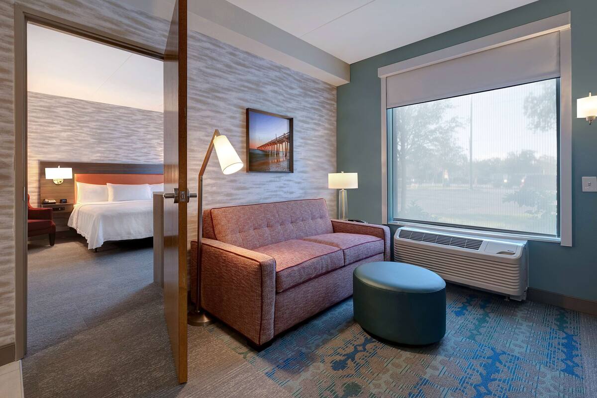 Ett hotellrum med en säng, en soffa, en liten pall, ett stort fönster och en tavla som hänger på väggen.